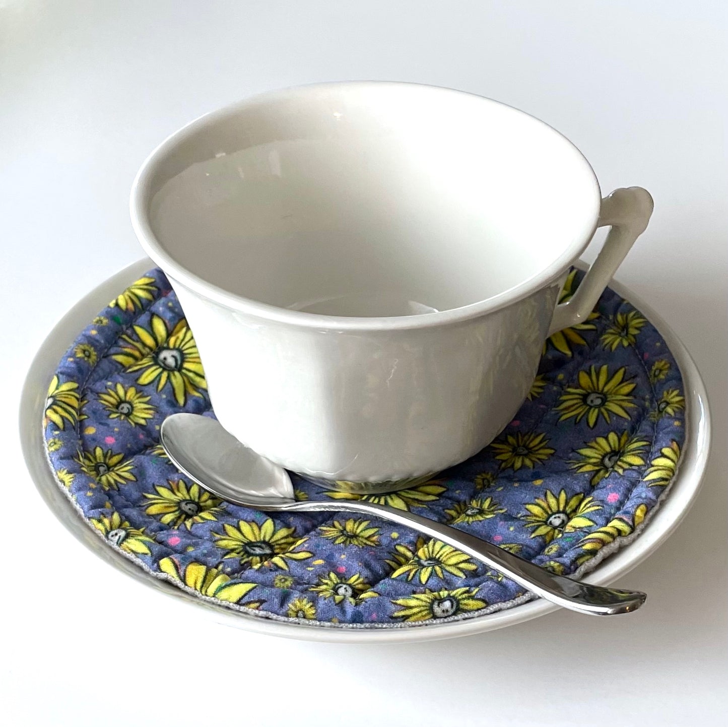 White teacup on top of purple saucer mat with yellow smiling flowers on white saucer.