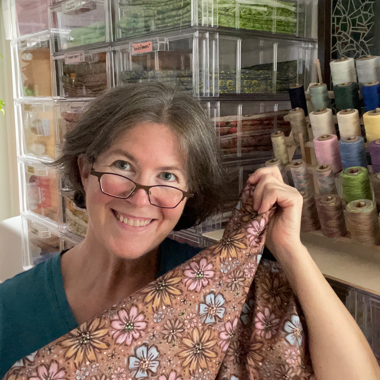 Kim holding brown floral fabric and smiling at the viewer, glasses pulled down making eye contact.