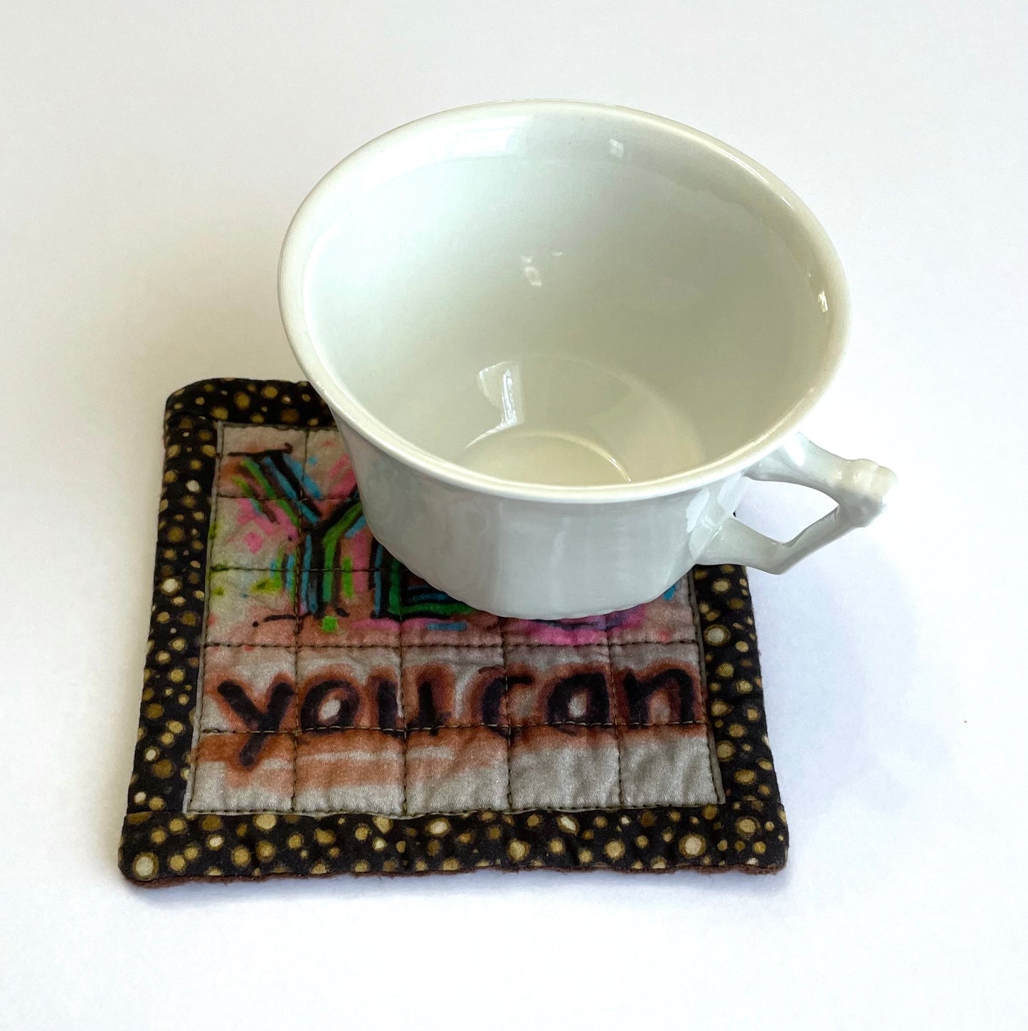 White teacup on top of square quilted mug mat coaster with Yes You Can design and brown polka dot border.