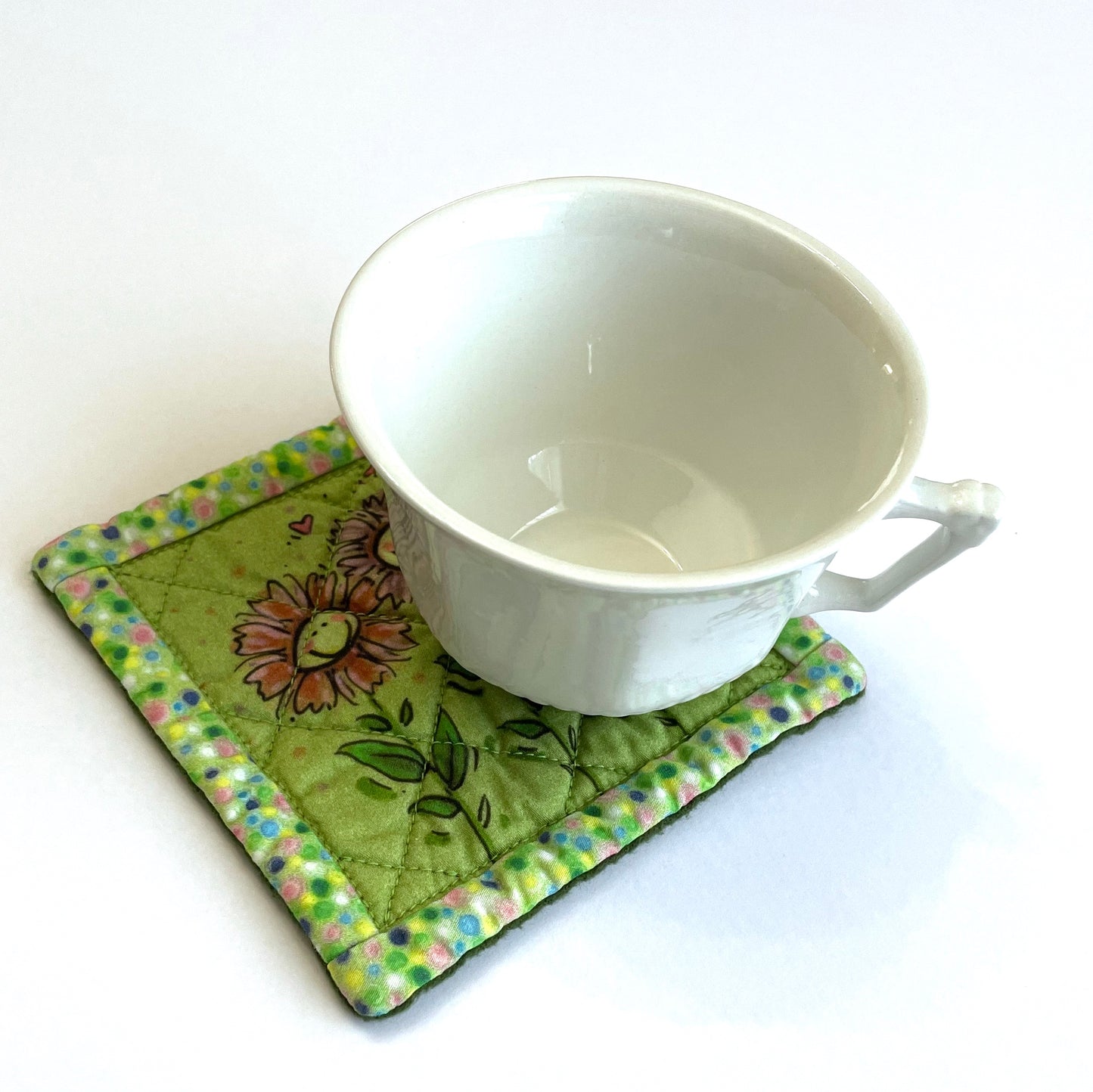 White teacup on top of square quilted mug mat coaster in avocado green with smiling flowers design and polka dot border.