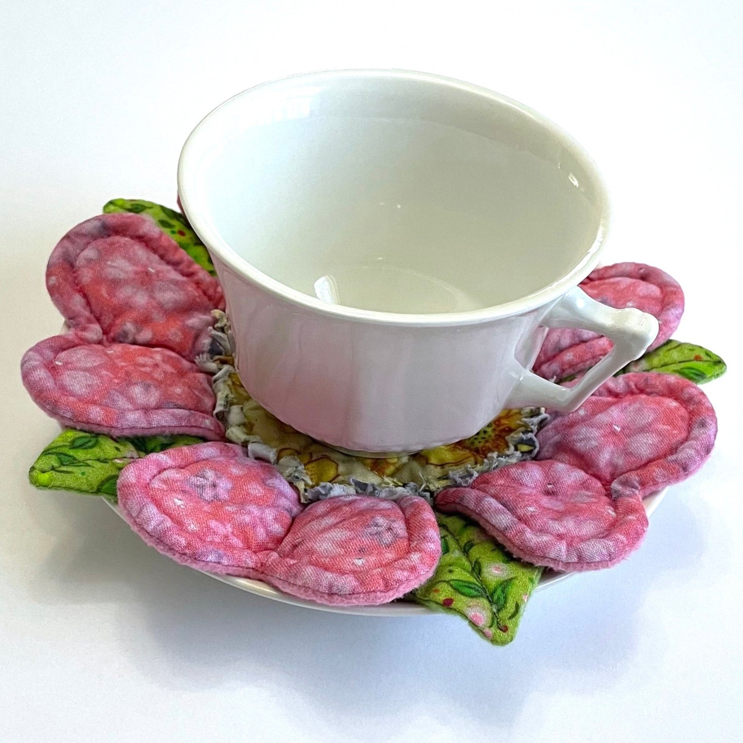 White teacup on top of quilted flower mug mat coaster with pink petals and green leaves. Cup and mug mat on top of small saucer.