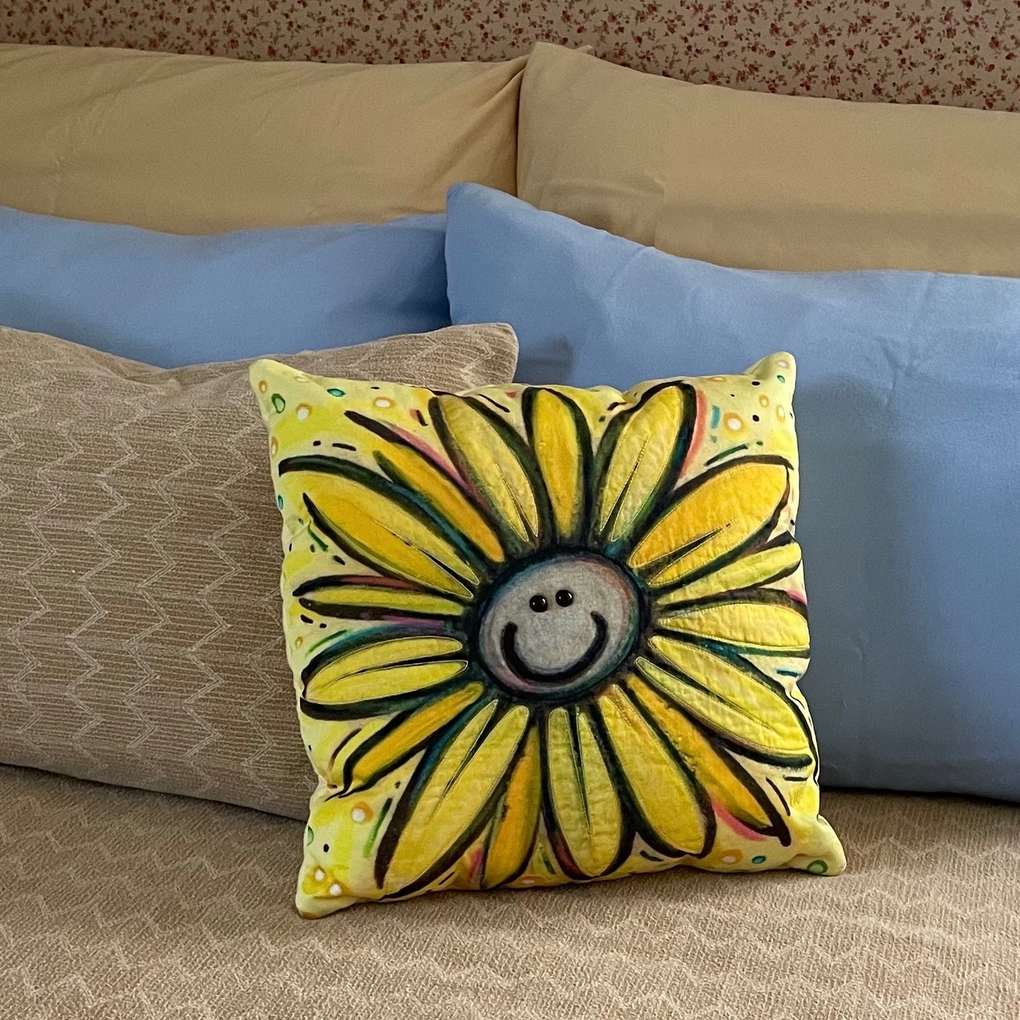 Bright yellow square throw pillow with big yellow smiley daisy design and shiny black eyes displayed on bed in front of background pillows.