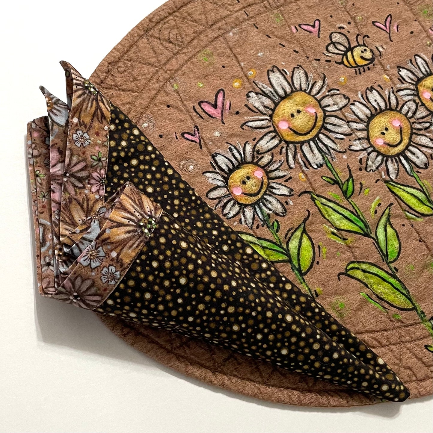 Quilted placemat oval smiling daisy design with cloth napkin in brown polka dots and rustic brown floral displaying curled triangle napkin fold.