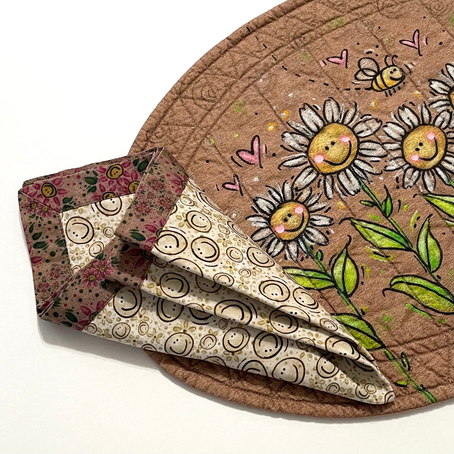 Quilted placemat oval daisies and bees with 2-color cloth napkin on top in triangle fold.