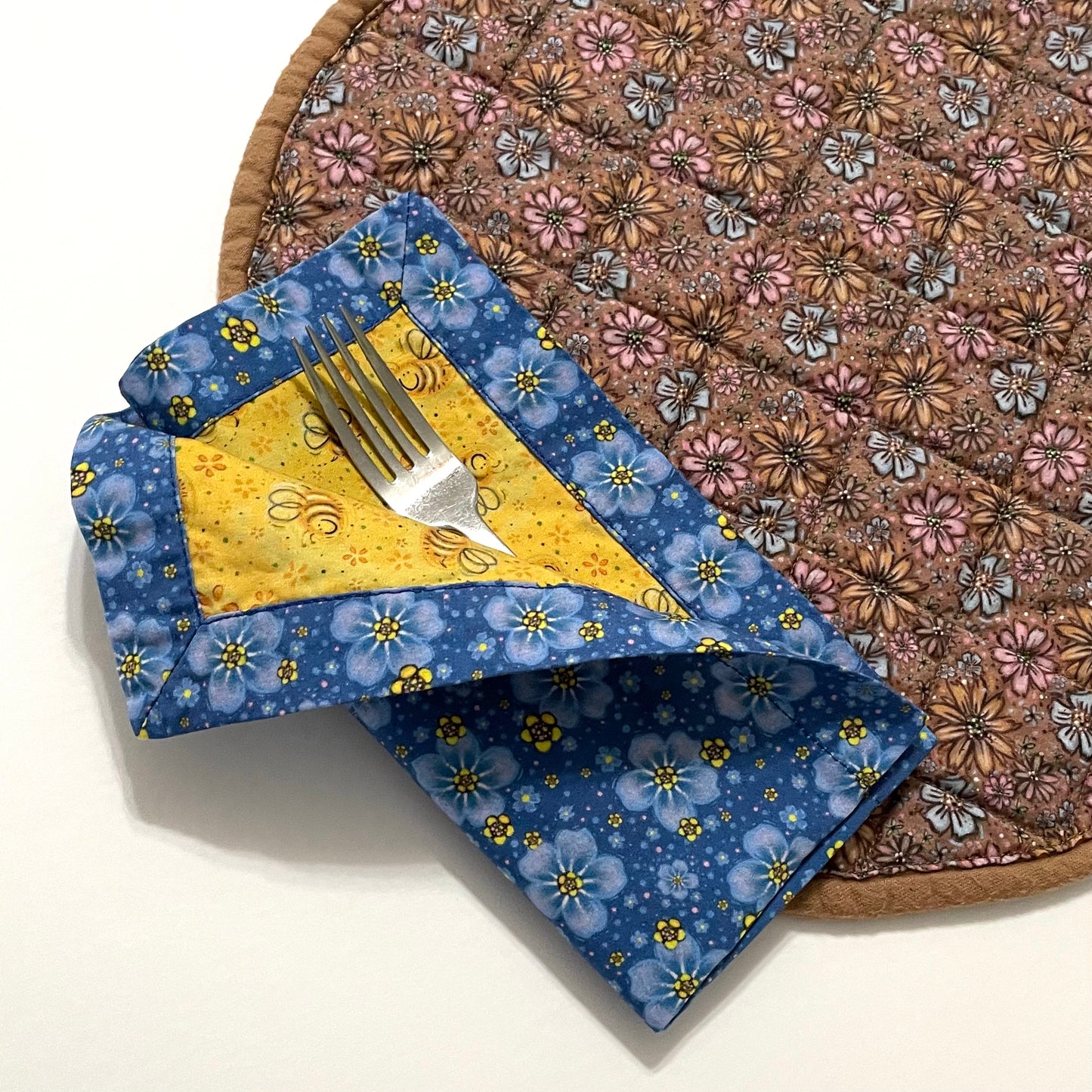 Quilted placemat round floral teatime style with reversible cloth napkin on top in yellow bees and blue floral displaying curled pocket napkin fold.