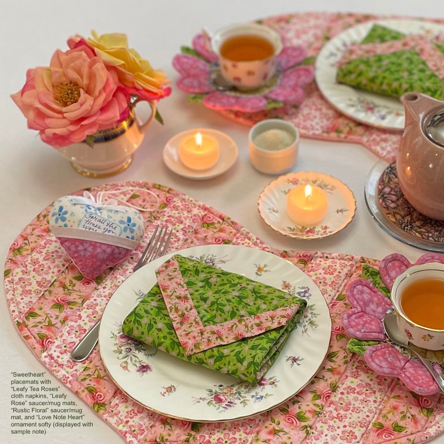 Romantic table setting with pink striped floral heart shaped placemats, envelope-folded cloth napkins on plates, heart shaped ornament softy gift, pink rose saucer mats on saucers with teacups, candles, and flowers in vase.